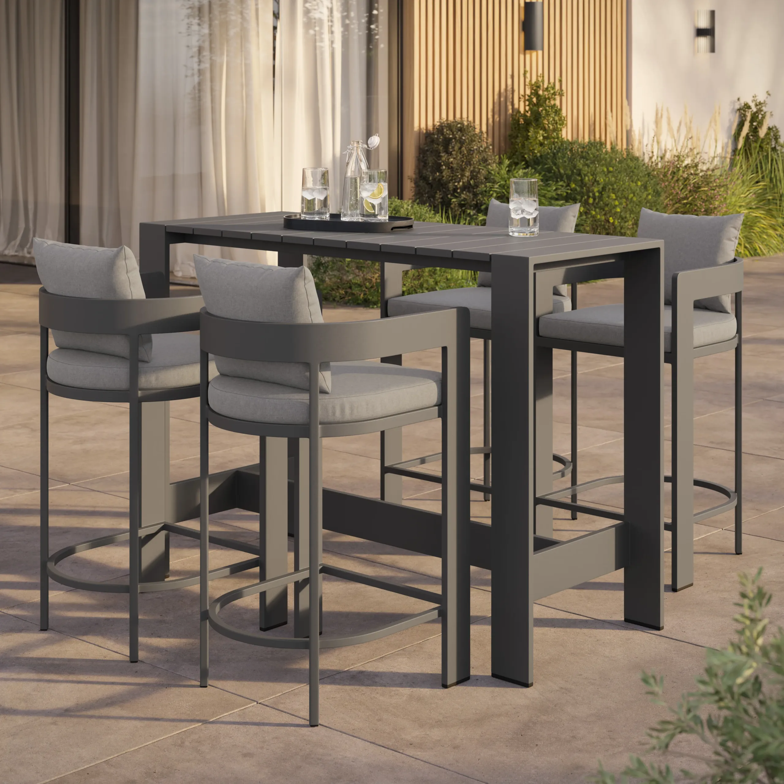 Modern dark-gray outdoor high dining table with four cushioned bar stools on a stone patio, plants and curtains behind.