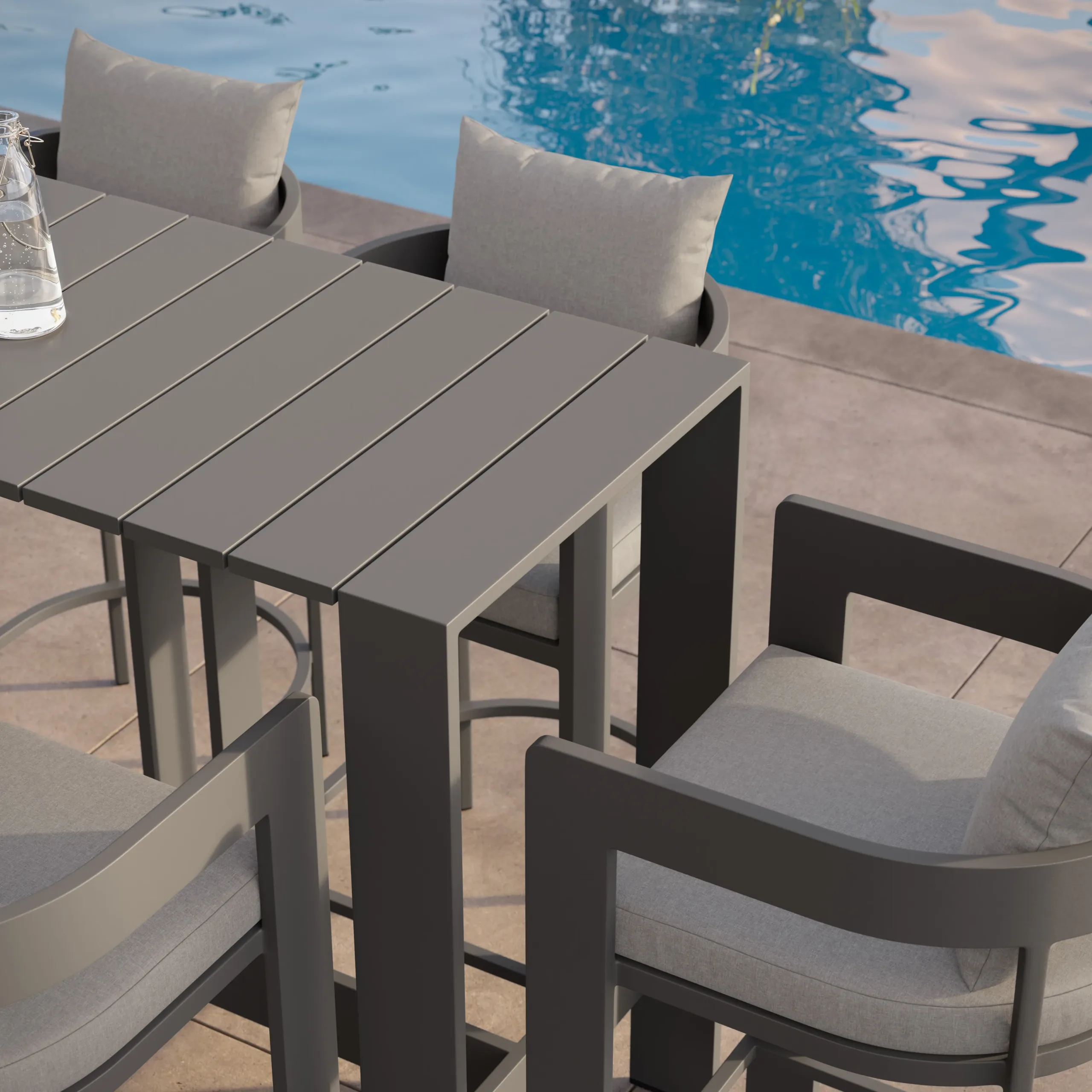 Gray slatted outdoor bar table with cushioned gray armchairs on a poolside patio with blue pool water.