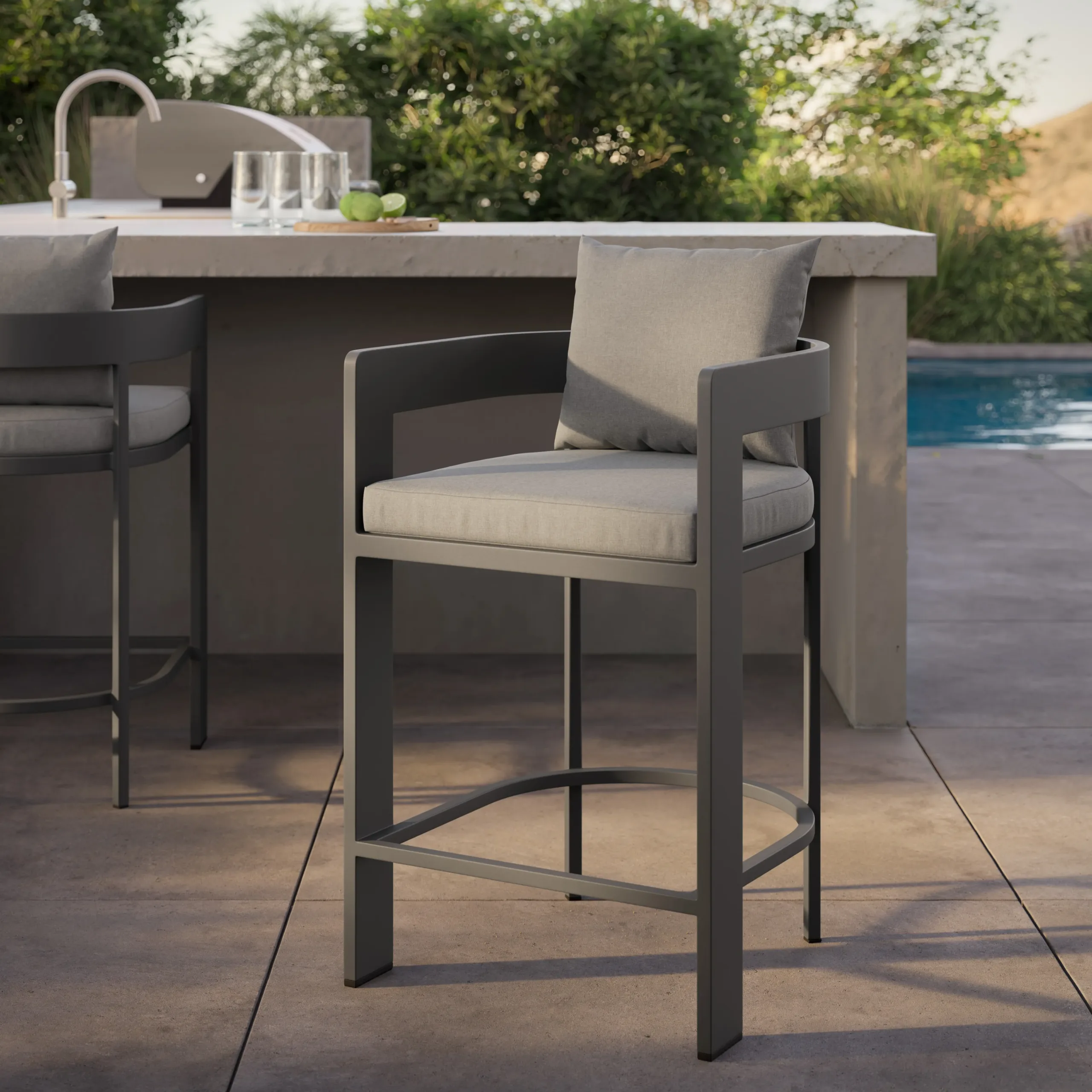 Modern gray metal outdoor barstool with light gray cushion and pillow, curved footrest, beside a patio bar and pool.