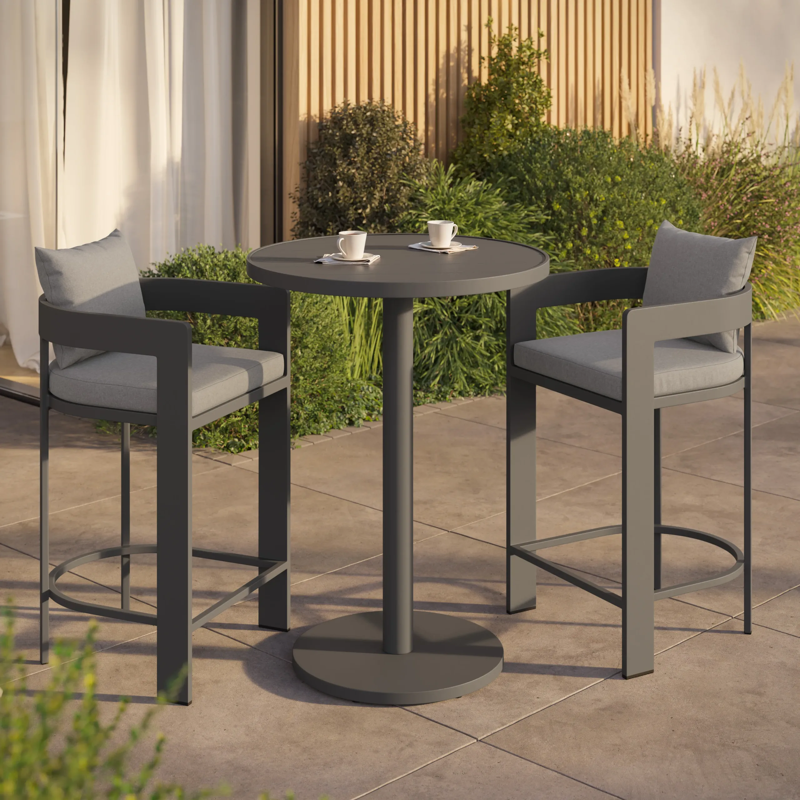 Round gray pedestal patio table with two matching high cushioned chairs and two white coffee cups, plants behind.