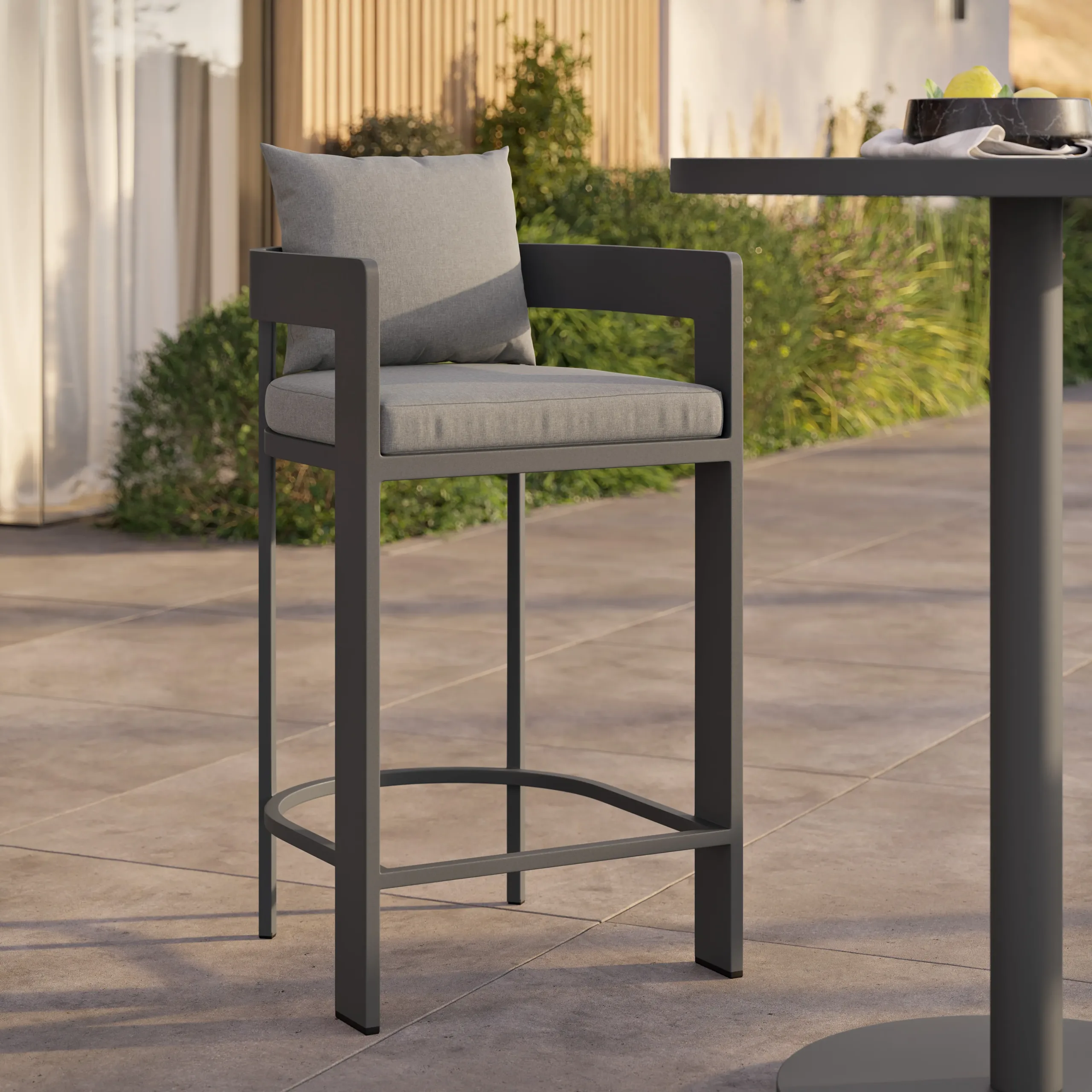 Modern gray metal outdoor bar stool with light-gray seat cushion and back pillow, curved footrest on tiled patio.