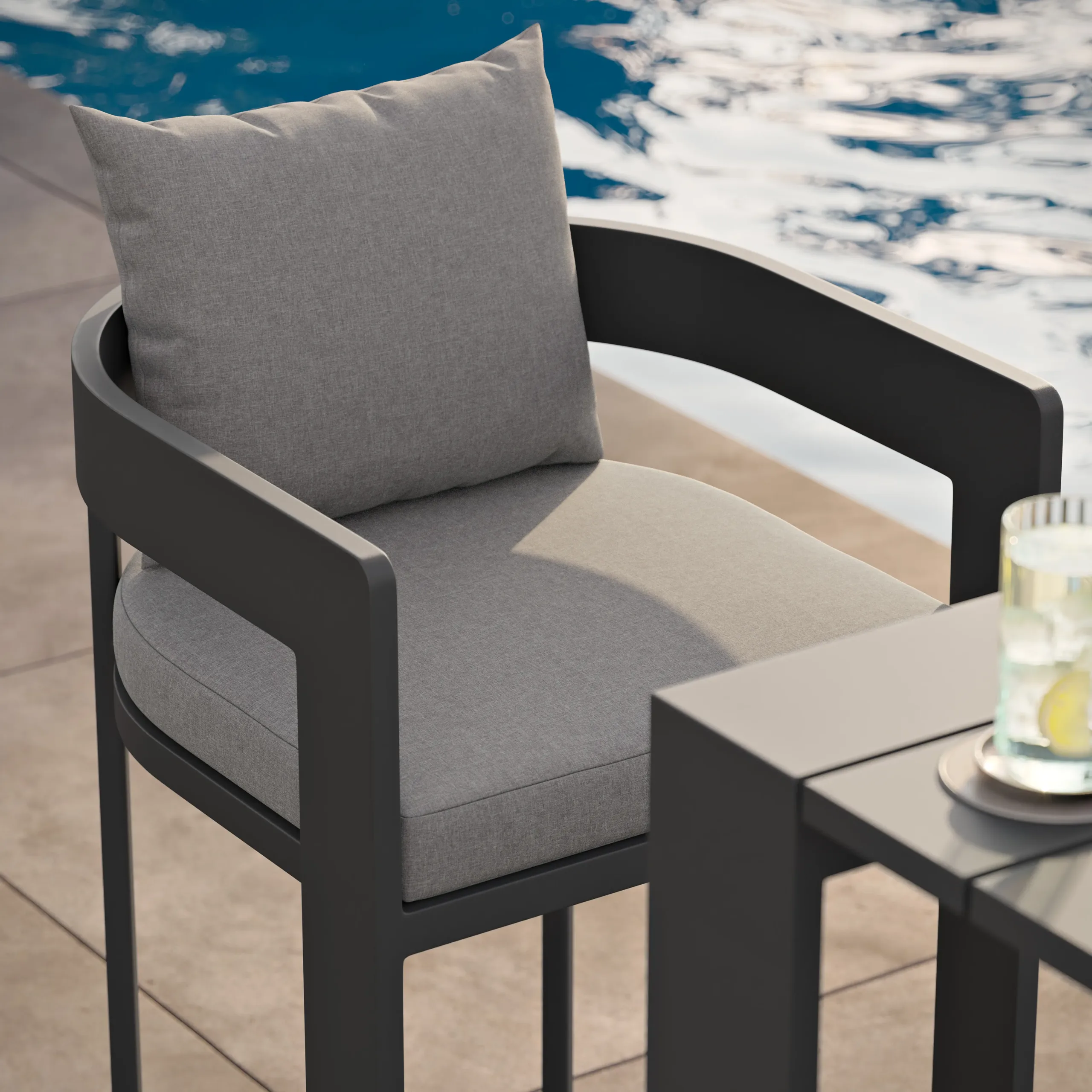 Gray cushioned outdoor armchair with dark metal frame beside a pool, next to a table holding a glass of lemon water.